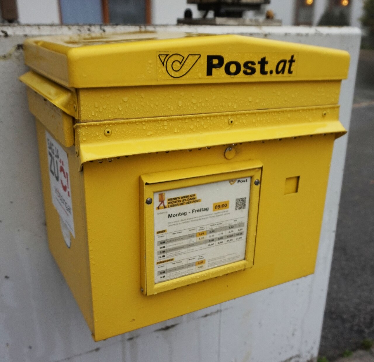 Gelber Briefkasten der Österreichischen Post mit Posthorn-Logo und Post.at-Schriftzug, regennass an einer weißen Wand montiert