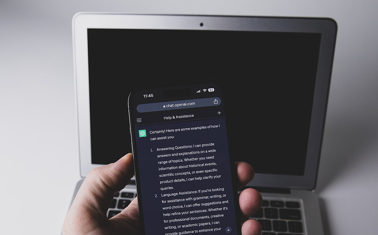 Hand hält ein Smartphone mit einer geöffneten Chatbot-Unterhaltung vor einem aufgeklappten Laptop