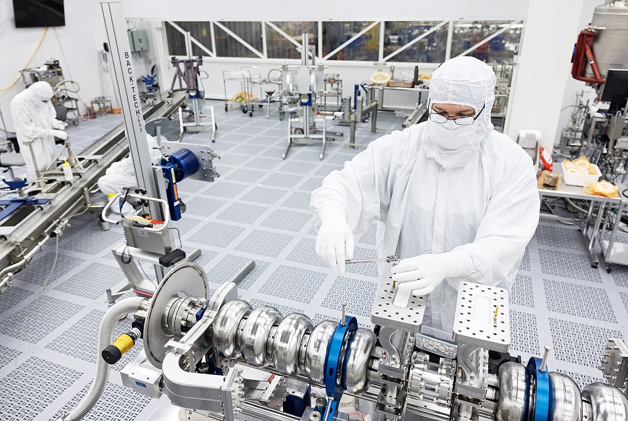 Techniker im weißen Reinraumanzug montiert Bauteile an einer Hightech-Anlage, im Hintergrund zwei weitere Beschäftigte im Reinraum