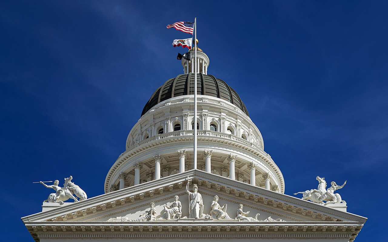 Die weiße Kuppel des kalifornischen Kapitols in Sacramento vor blauem Himmel, darüber die US- und die Kalifornien-Flagge